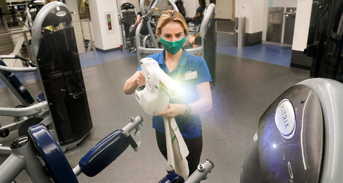 Student worker sprays disinfectant on gym equipment.