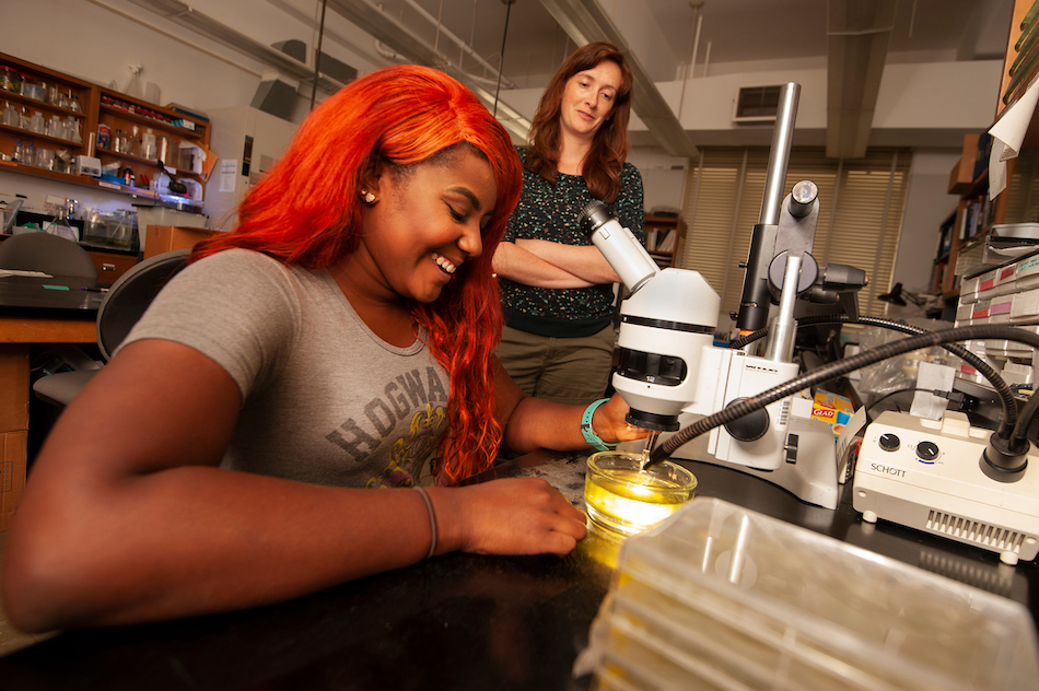 A student smiles as she inspects yellow liquid underneath a microscope, while her professor watches on.