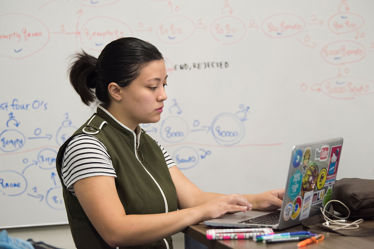 A student works on her laptop in front of a whiteboard with a complex math equation.