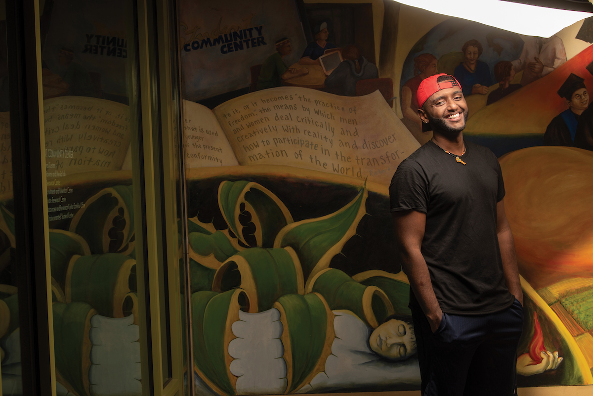 A smiling sociology major in a red baseball cap stands in front of a colorful mural.