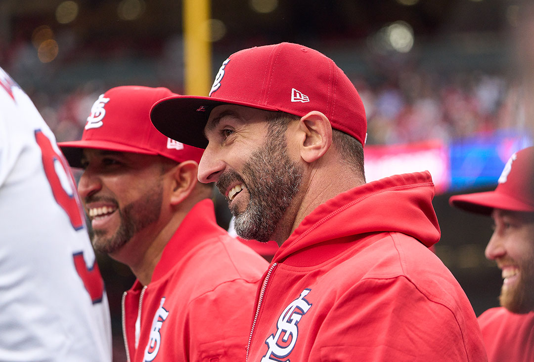 Descalso-Cardinal-Coach A man in a red cap and jacket smiles looks off camera, smiling. He is in a dugout with other people wearing red.