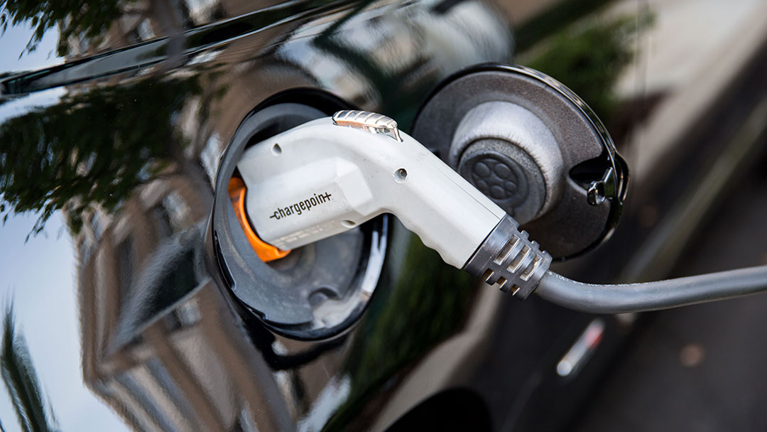electric-vehicle-charging A closeup photo of a car being charged. We see a cap on the side of the vehicle open, with a white charger plugged in.