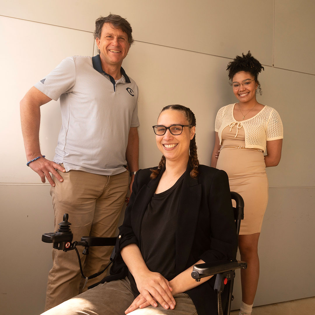gregory-downs-chantal-walker-serena-crawford Three people pose for the camera. The person on the left stands with a hand on their hip, while a second sits in a wheelchair and a third stands to the right, hands behind their back.