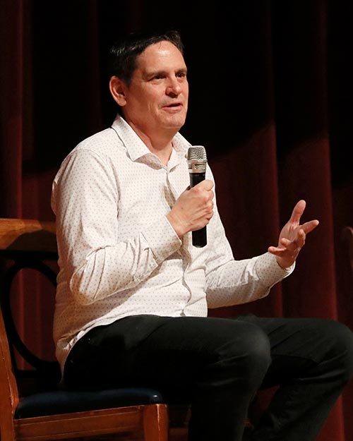 mejdal-inset A man in a white shirt sits on stage, holding a microphone and talking.