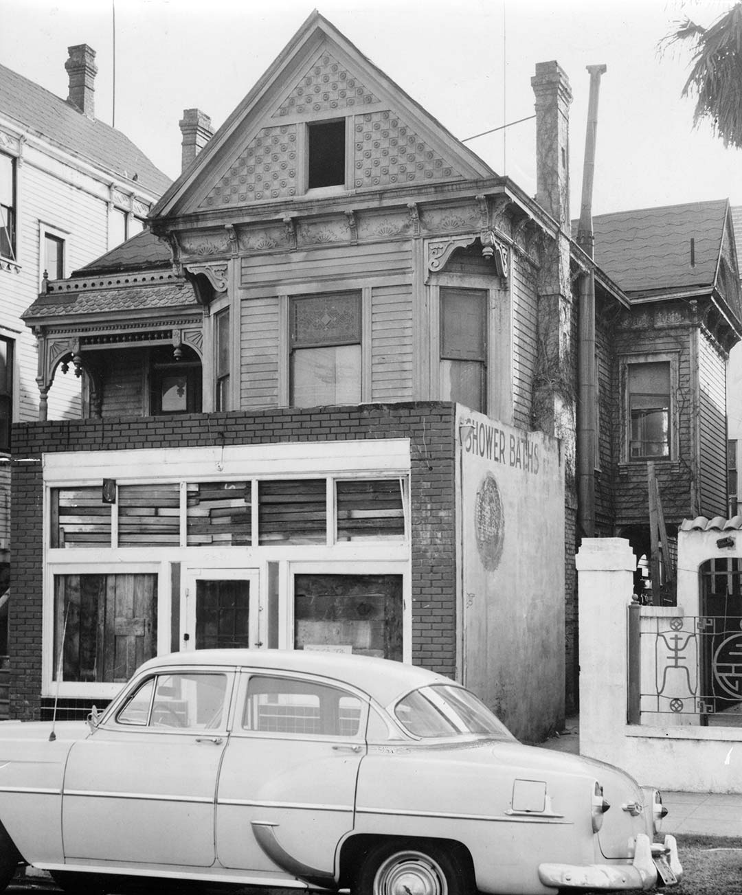 redevelopment-house-demo Black and white photo of a three story house. The house has a peaked roof, bay window in the front on the second floor and a closeed-in porch on the ground floor. A sedan is parked on the street in front. The windows are boarded up.