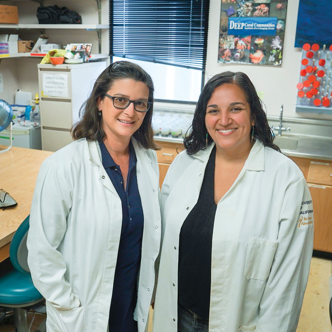 women-mentors-oceans Two women scientists in lab coats stand together in a lab, facing us and smiling.