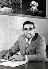 Black and white photo of a white man with dark hair wearing a jacket and tie sitting at a desk. Above and behind is head is a photo of a mushroom cloud.
