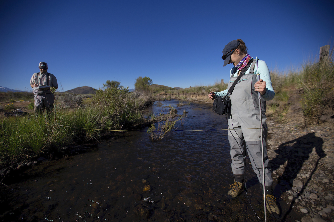 Woman scientist stands in stream with monitoring equipment with male scientist in background
