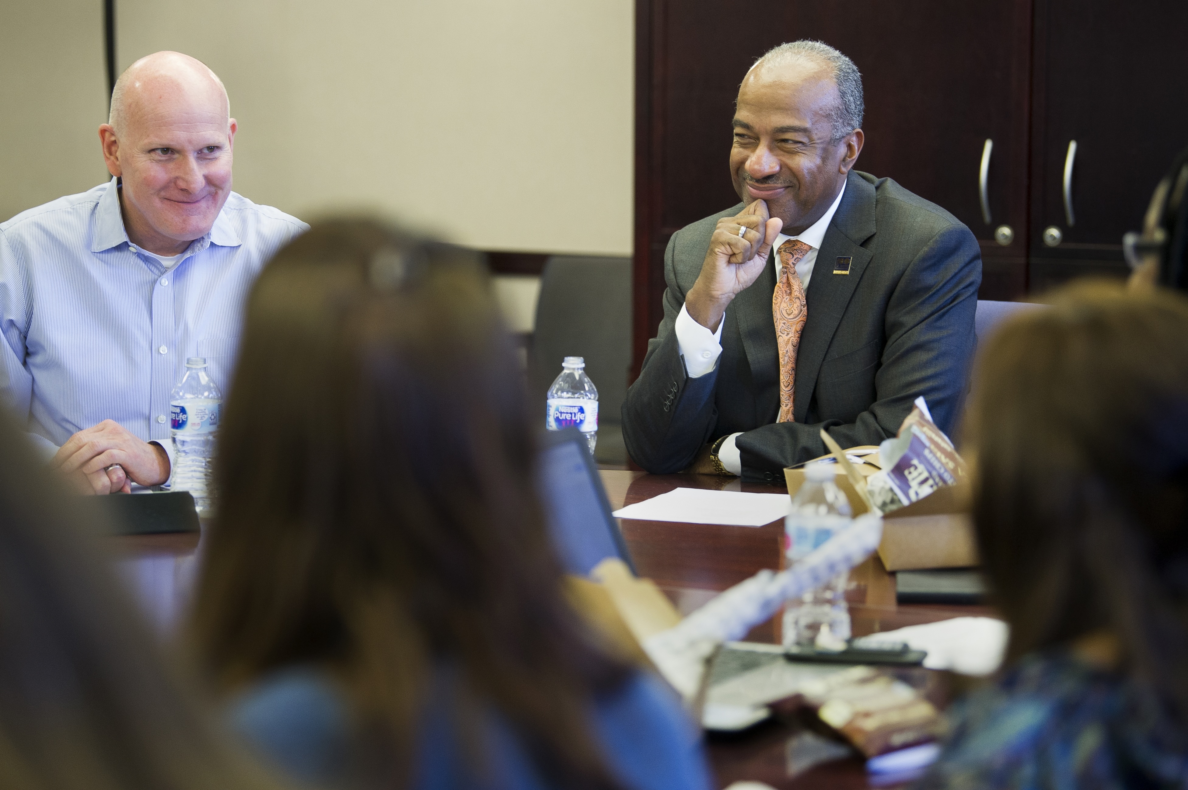 Two men smiling at a conference table, photo; blurred attendees in foreground