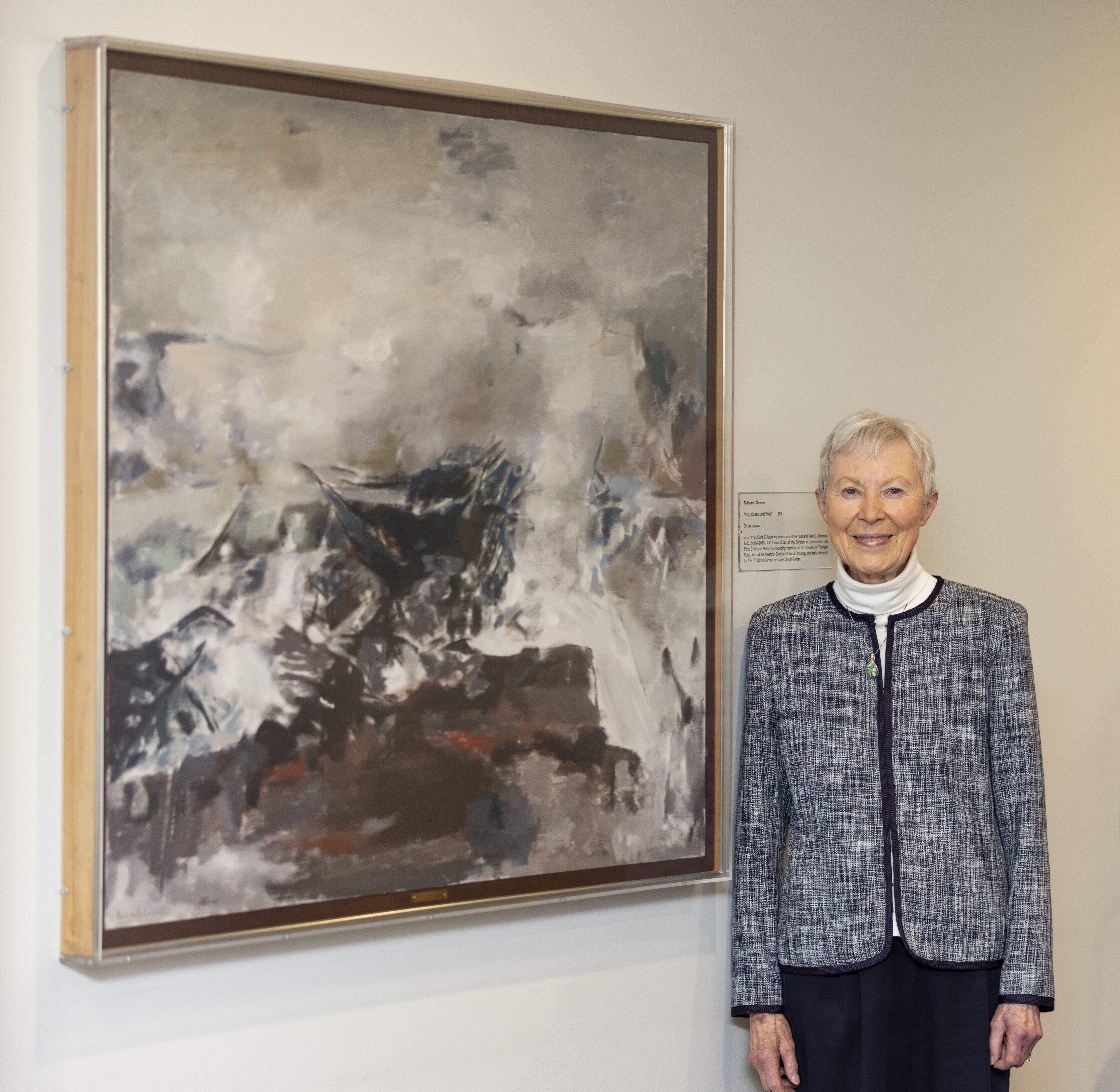 A woman with short white hair stands to the right of an abstract painting of white and blue waves crashing on a rugged shoreline.
