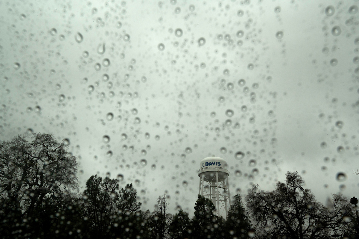 landscape of UC Davis water tower as seen through rain-splattered window on a gray day with above a silhouette of trees.