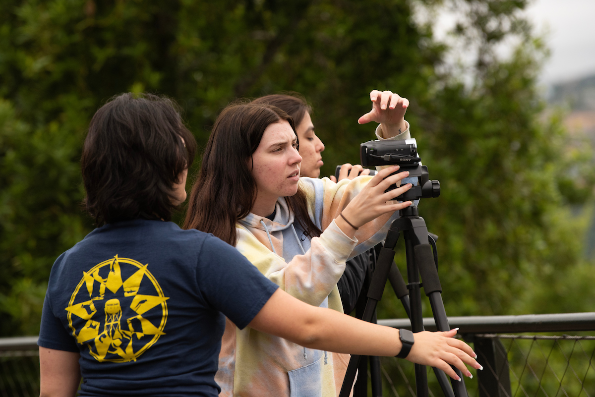 Three students look through a telescope on a platform surrounded by trees.