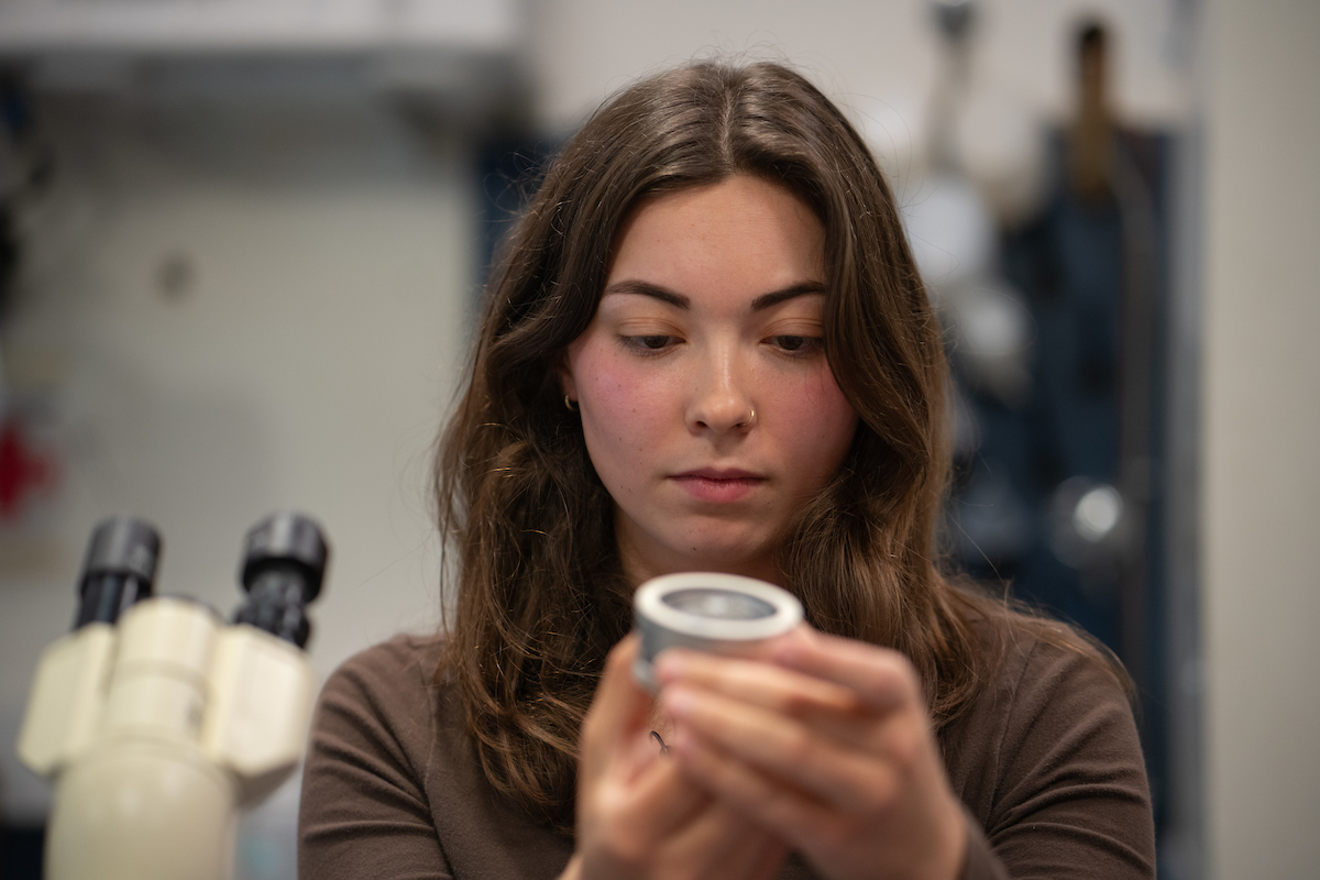 Sarah Howe in brown shirt and brown hair holds circular device containing an otolith close to her face as she looks at it