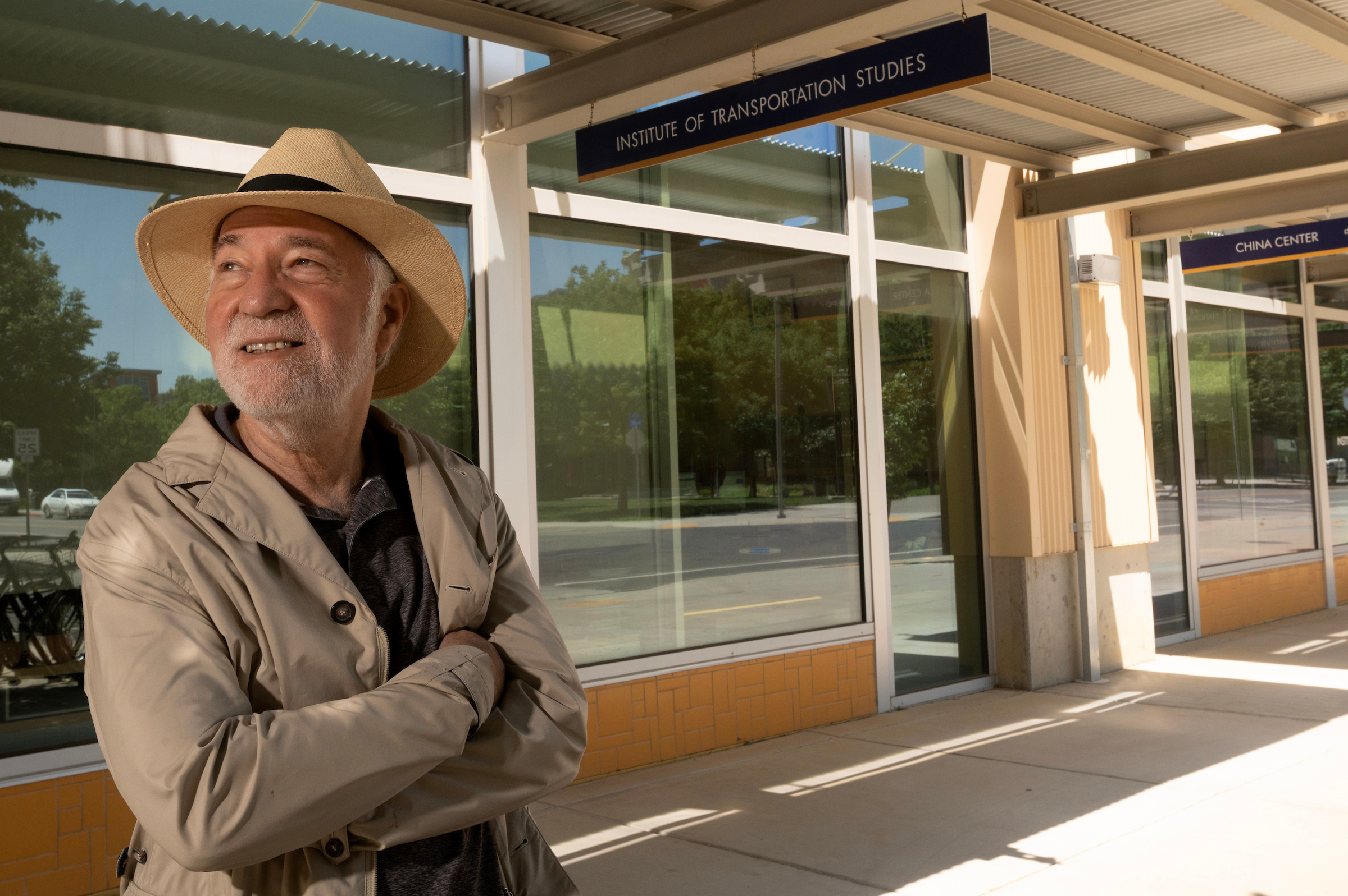 Outside a university building underneath a sign that reads "Institute for Transportation Studies" stands a man in a beige fedora hat and jacket, calmly looking to their right, with their arms folded across their chest.