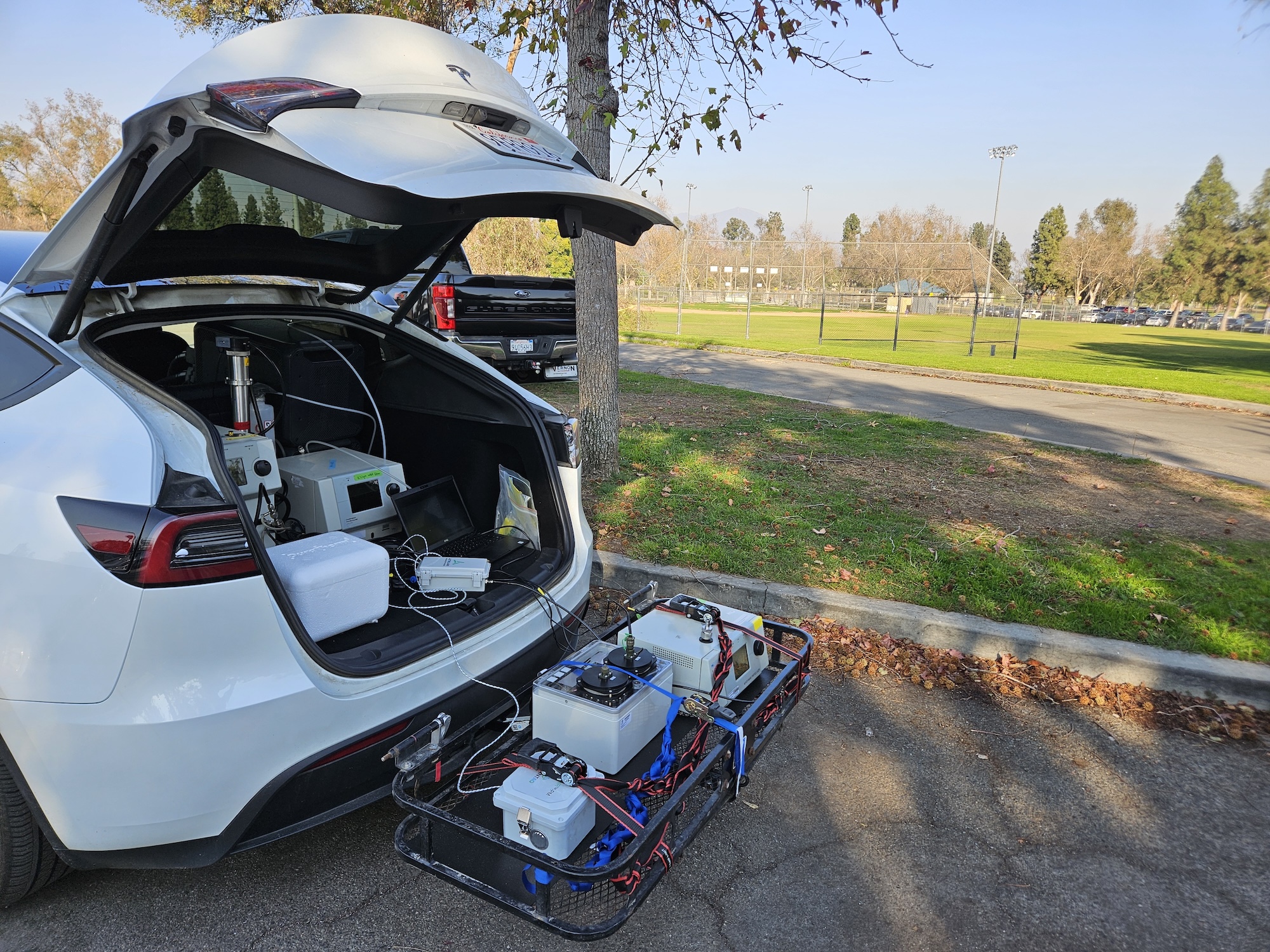 White car with open trunk holds air sampling and monitoring equipment. Background is green grass and tree.
