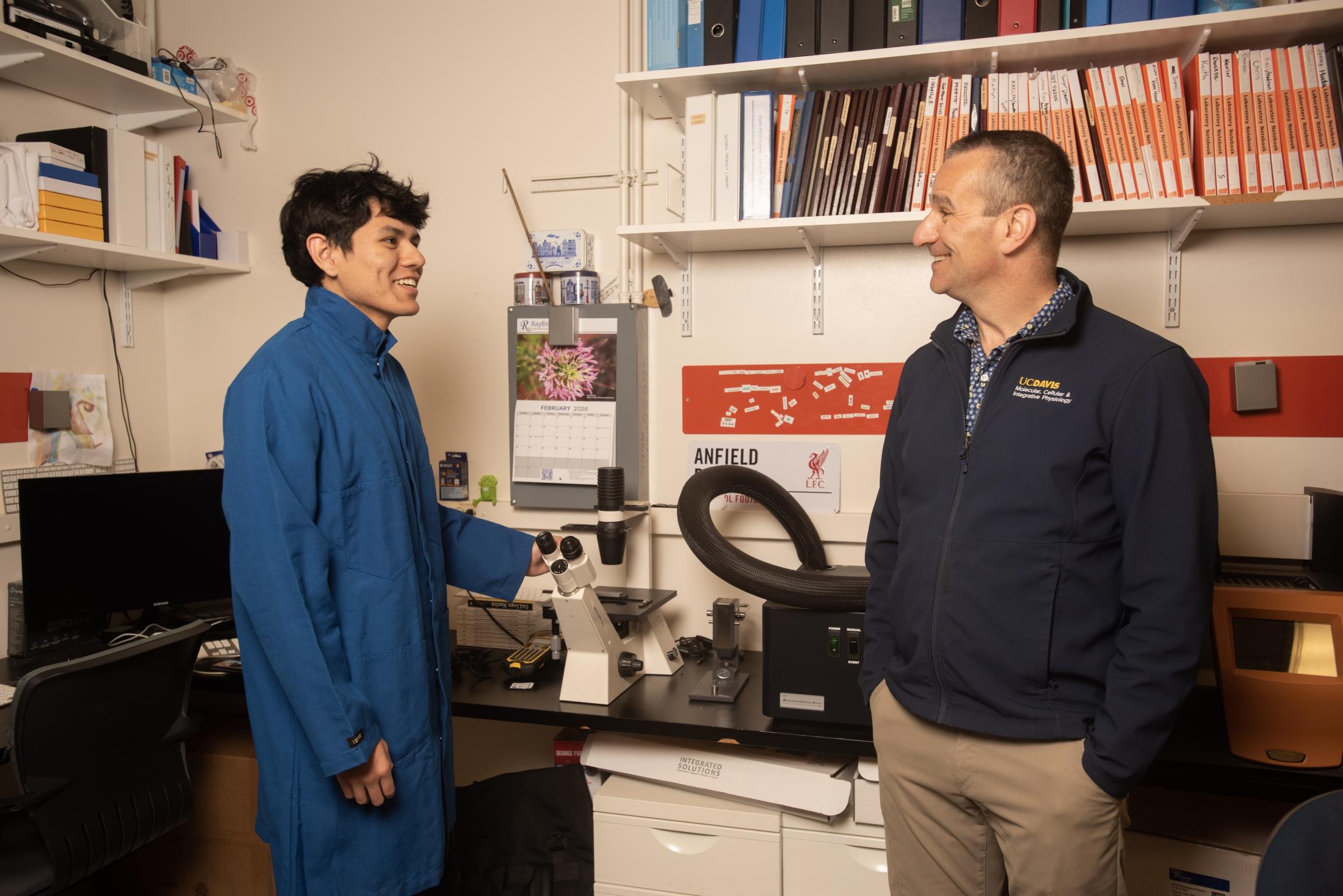 Undergraduate researcher in a blue lab coat smiles while speaking with a faculty member beside a microscope and imaging equipment inside a campus laboratory office in Briggs Hall.