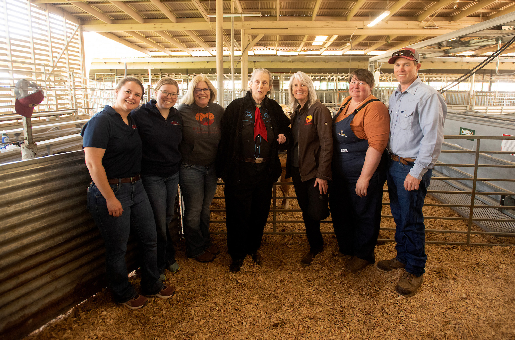 Seven people pose together inside a barn, smiling in front of animal pens. Left to right: Ashlynn Kirk, Karly Anderson, Cassandra Tucker, Temple Grandin, Jen Walker, Fauna Smith, Nathan Medlar. (Gregory Urquiaga / UC Davis)
