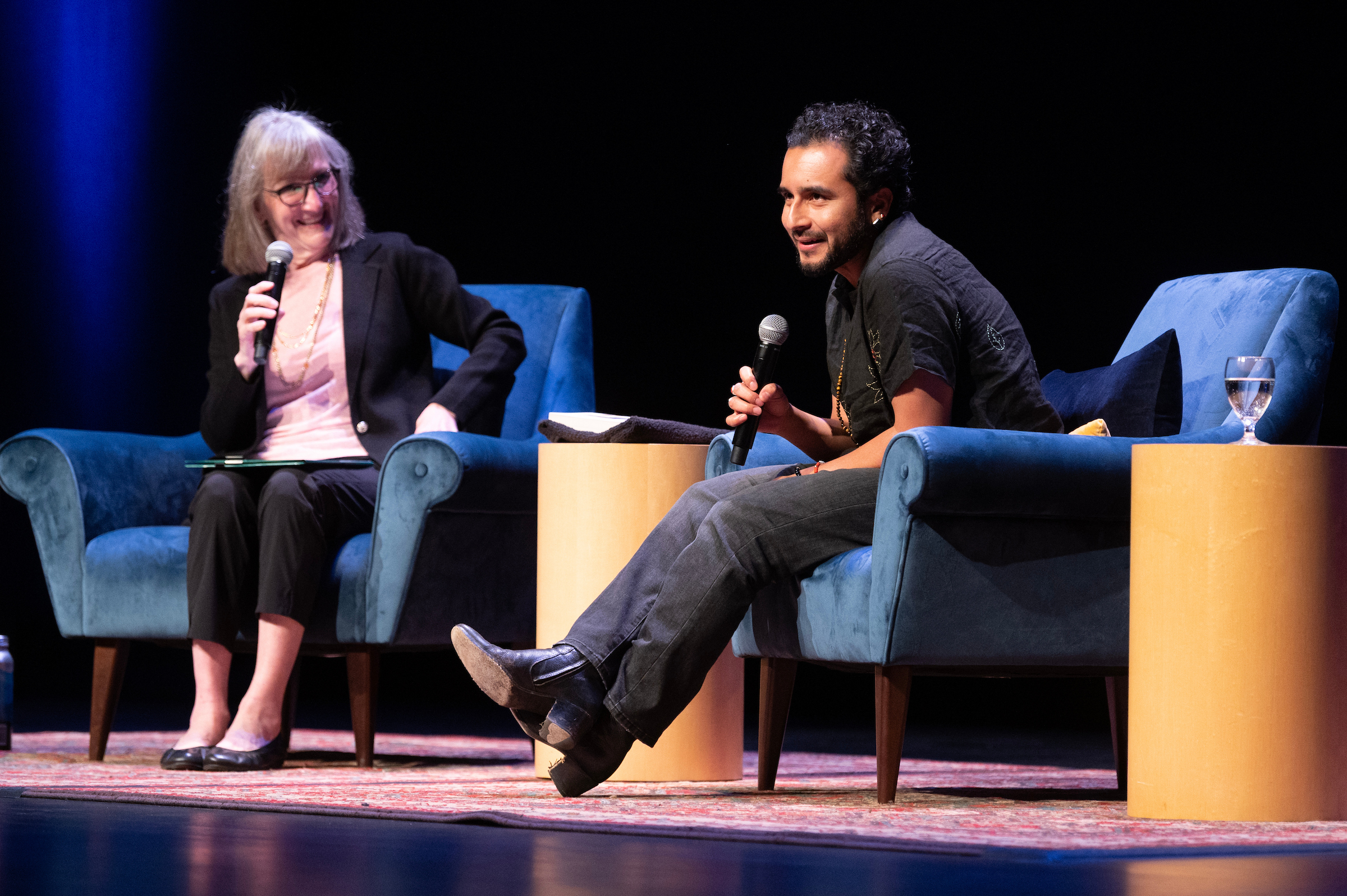 Two presenters -- Donna Apidone to the left and Javier Zamora to the right -- sit on a stage in blue chairs, engaging during a discussion.