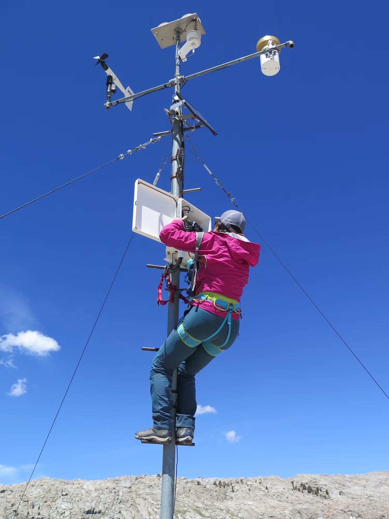 MJ Farruggia in pink jacket and safety harness climbing a weather station pole against blue sky.