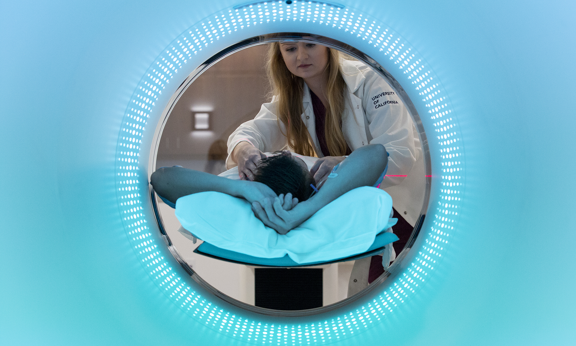 In a circular opening, a long-haired woman in a white lab coat bends over a person lying down with the top of their head towards the camera. Blue color fills the rest of the field outside the circle.