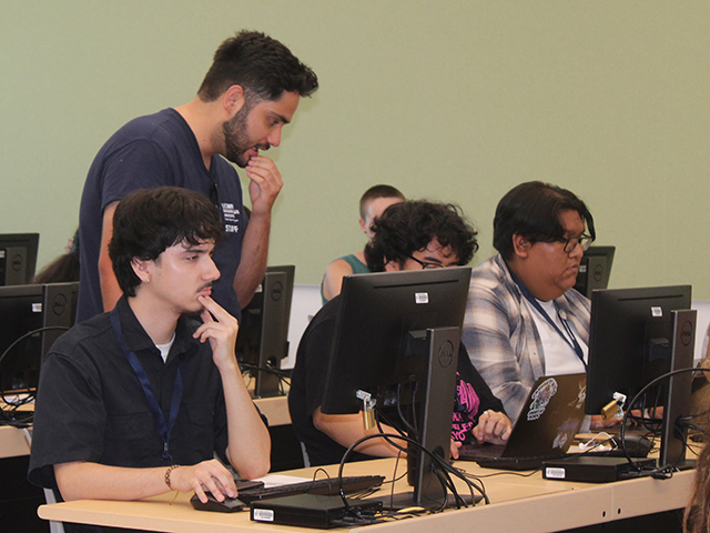 Instructor leans over students working at desktop computers in a classroom.