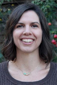 Smiling woman with medium-length brown hair, wearing a sweater, in a garden setting.