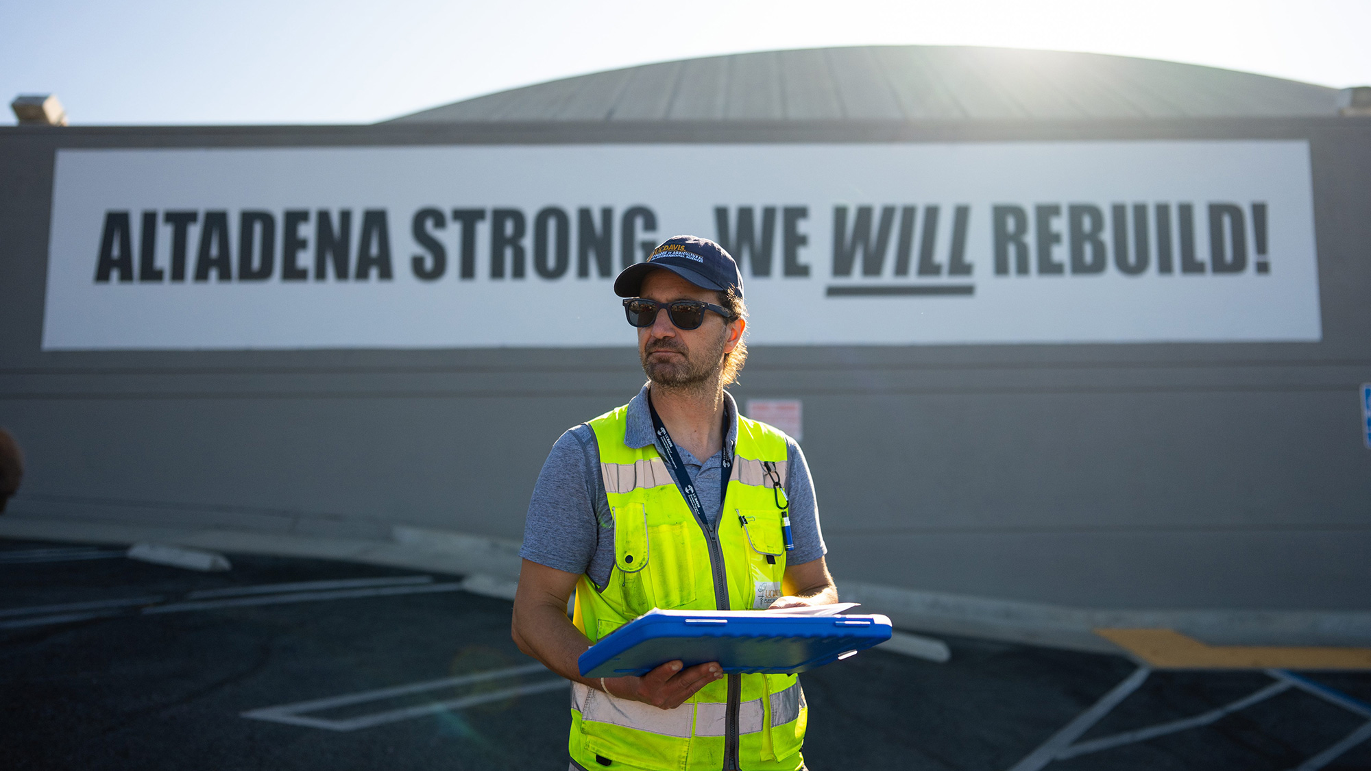 Man in a cap and vest holding clipboard beneath 'Altadena Strong' banner
