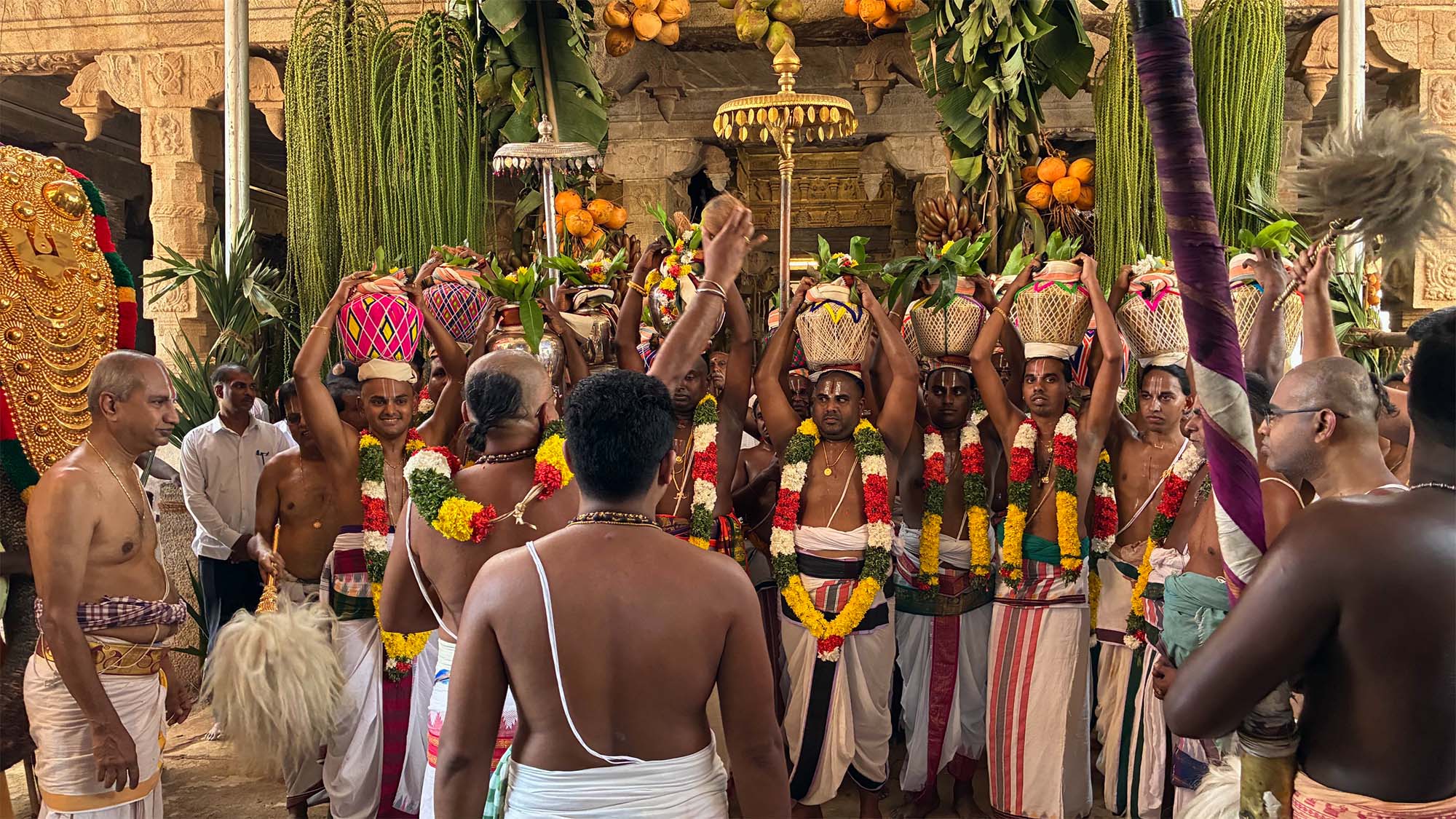 A group of men in traditional attire holding pot offerings during a ceremony in a decorated hall.
