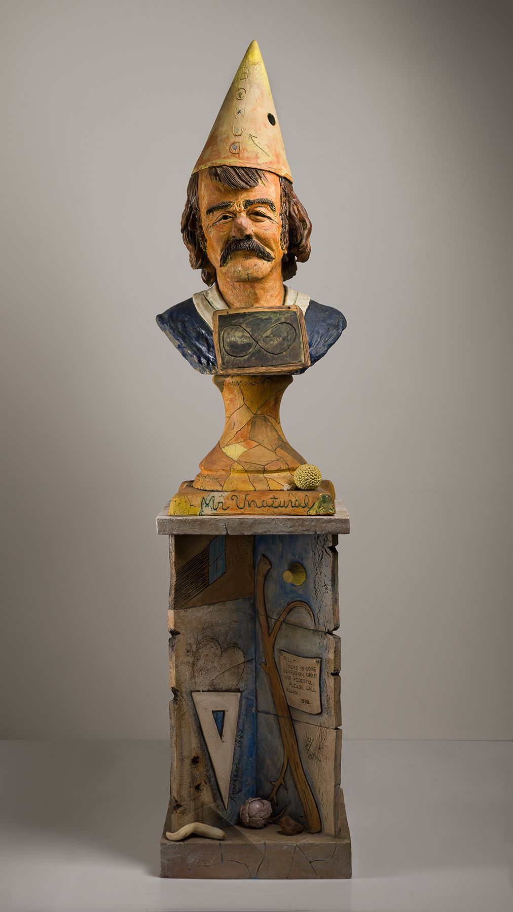 Weathered wooden bust with cone hat on painted cabinet containing found objects. 