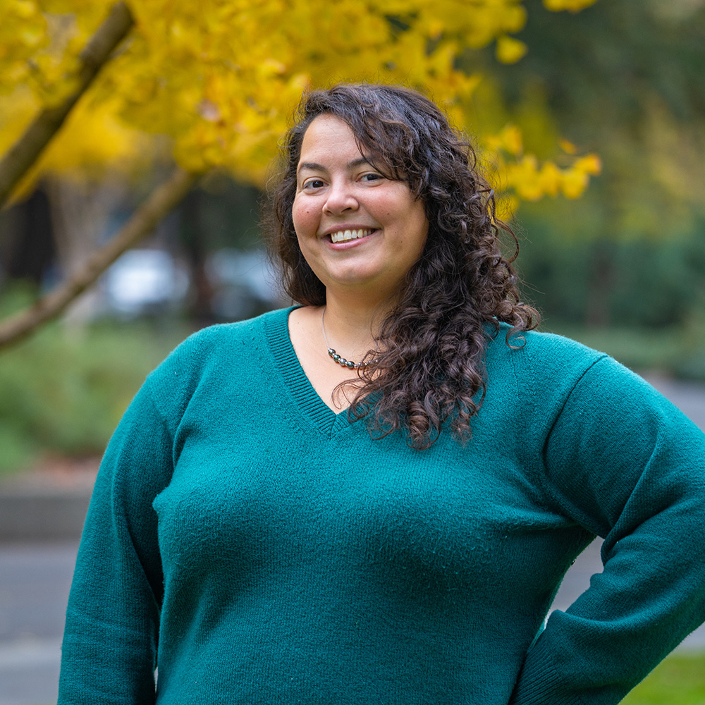 Smiling woman with curly hair in teal sweater, hand on hip, yellow autumn tree behind