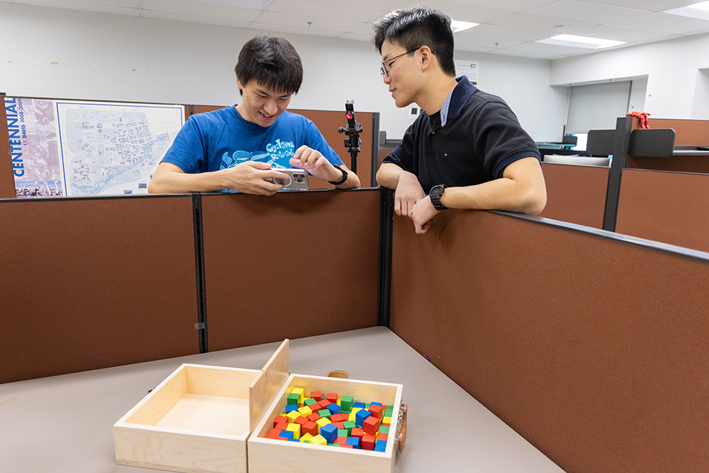 Two people leaning on office cubicles discussing while looking at a smartphone, with a box of colorful shapes on the table.