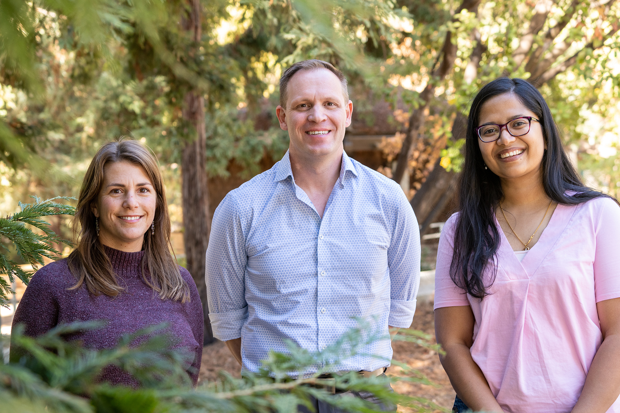 Three people outdoors facing the camera. From left, woman with shoulder length blonde hair, purple top; man with short hair, light blue shirt; woman with long black hair and glasses, pink top.