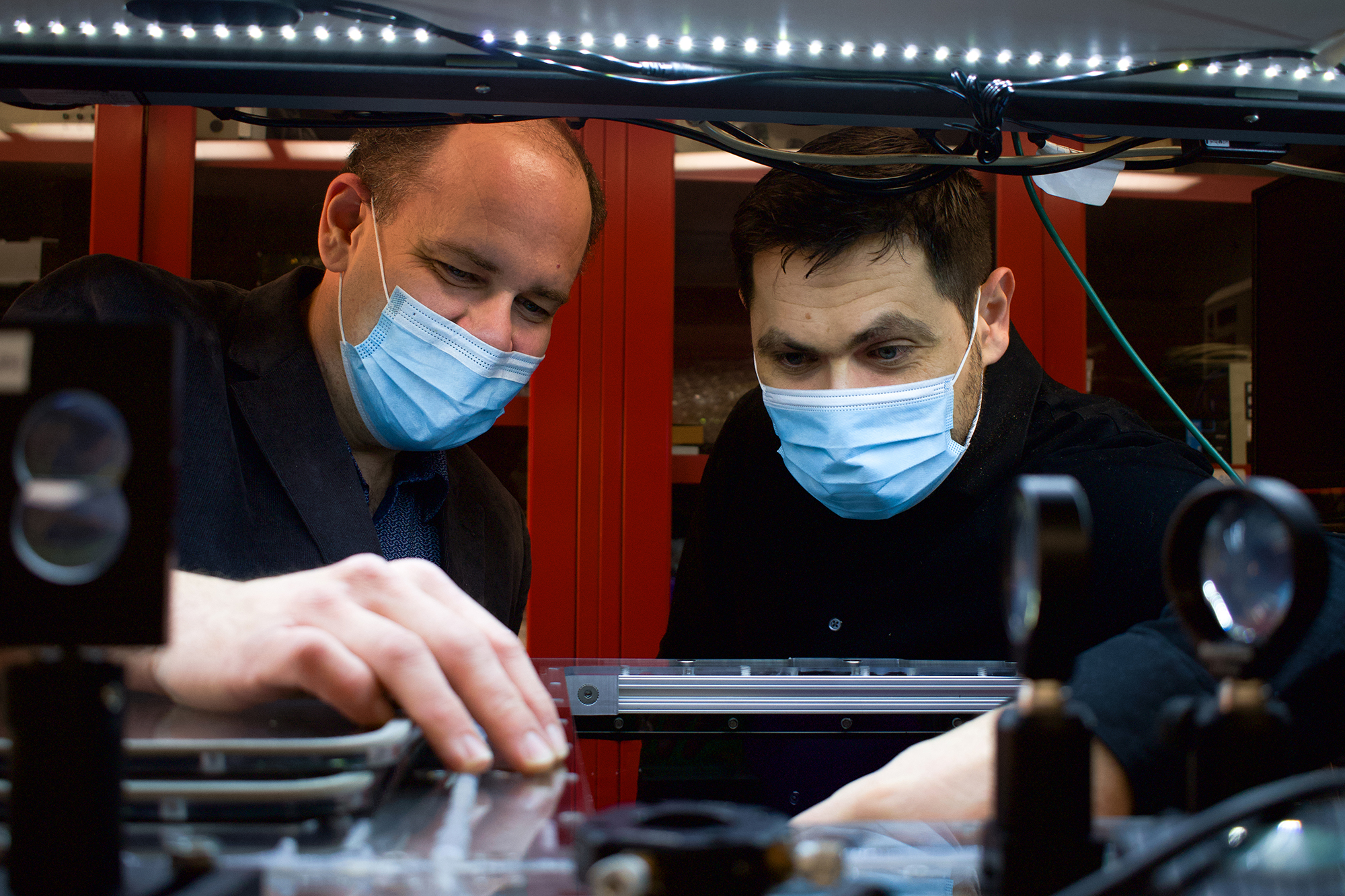 Two men in dark blue lab coats look at something on a lab bench. 