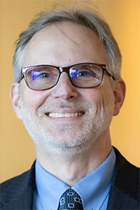 Smiling man with glasses and a suit, set against a warm, soft-focus background.