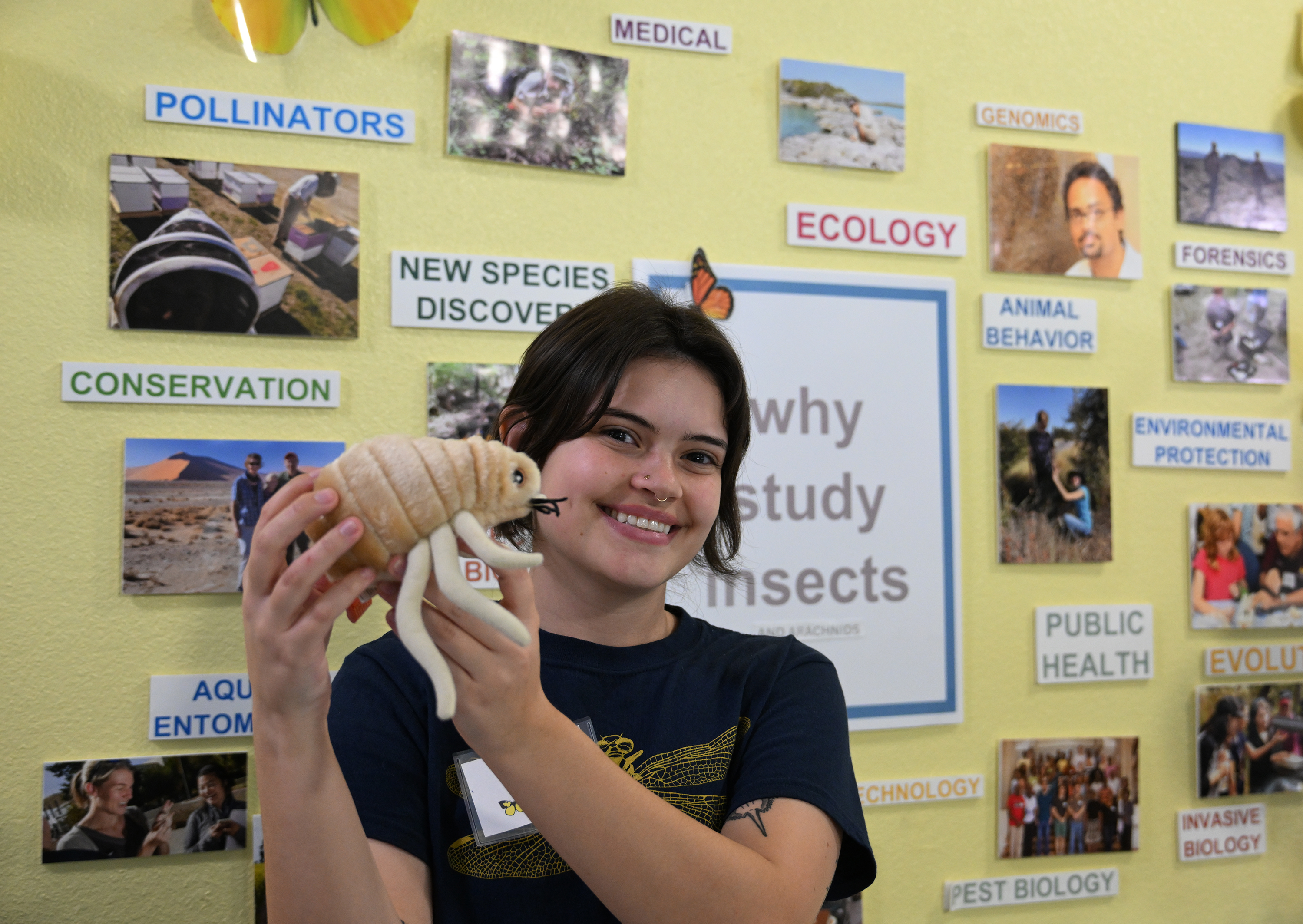 In a museum setting, a student upholds a stuffed animal in the shape of a tick, modeling the stuffed toy for the camera. 