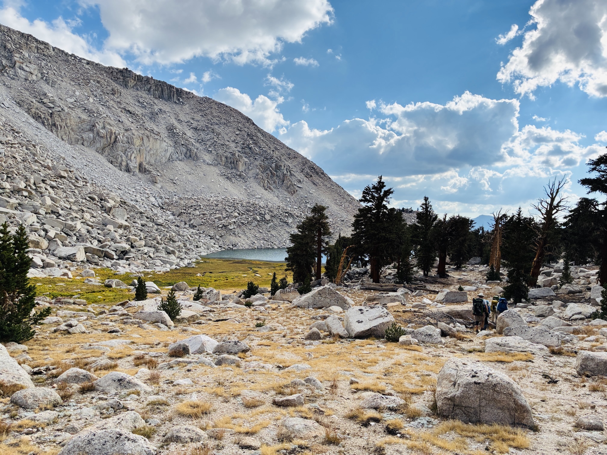 Hikers walk across Sierra Nevada landscape doted with a few dark green pine trees, mountain slope, deep yellow meadow, and lake