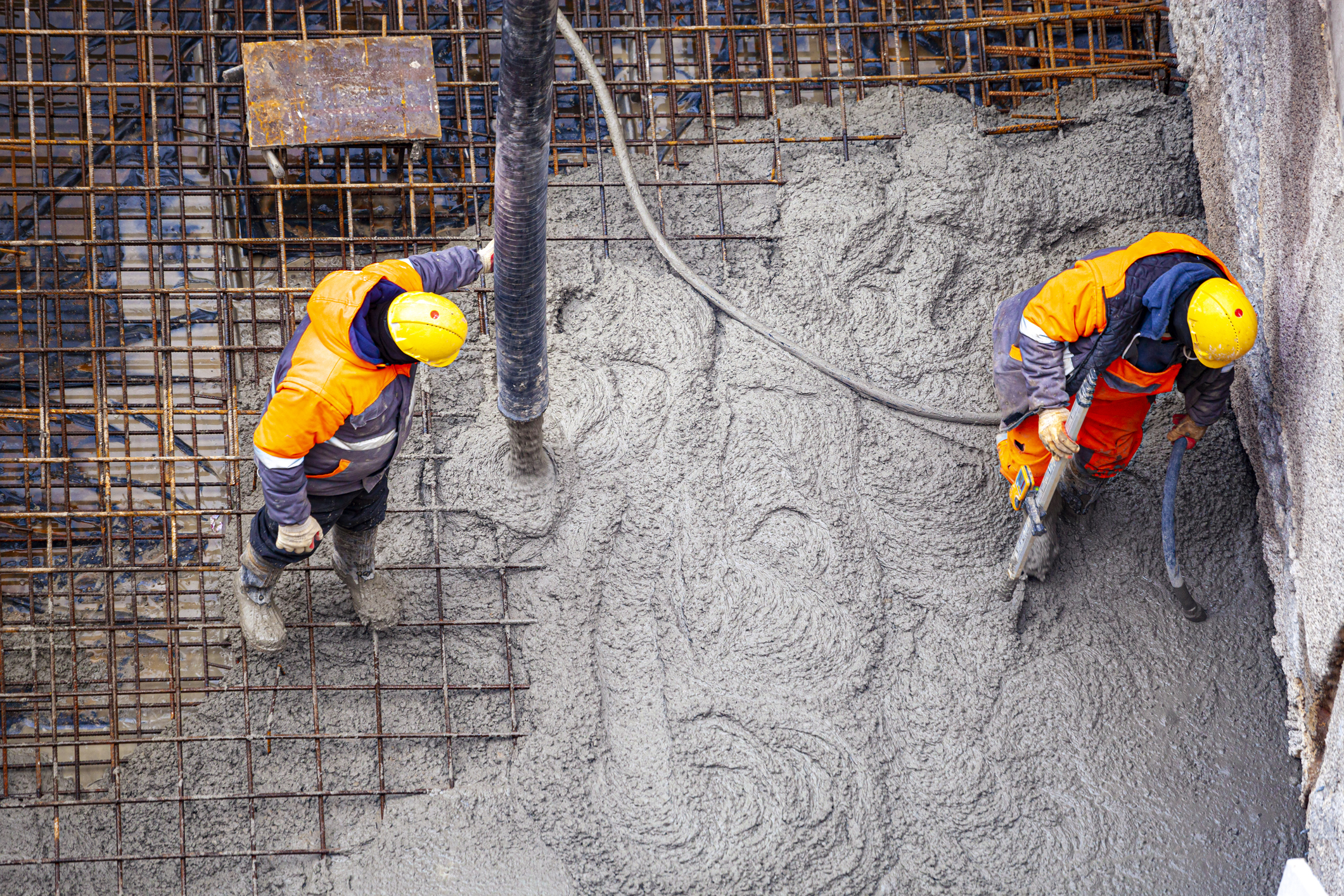 Aerial photo of two workers smoothing wet concrete among rebar wearing yellow helmets