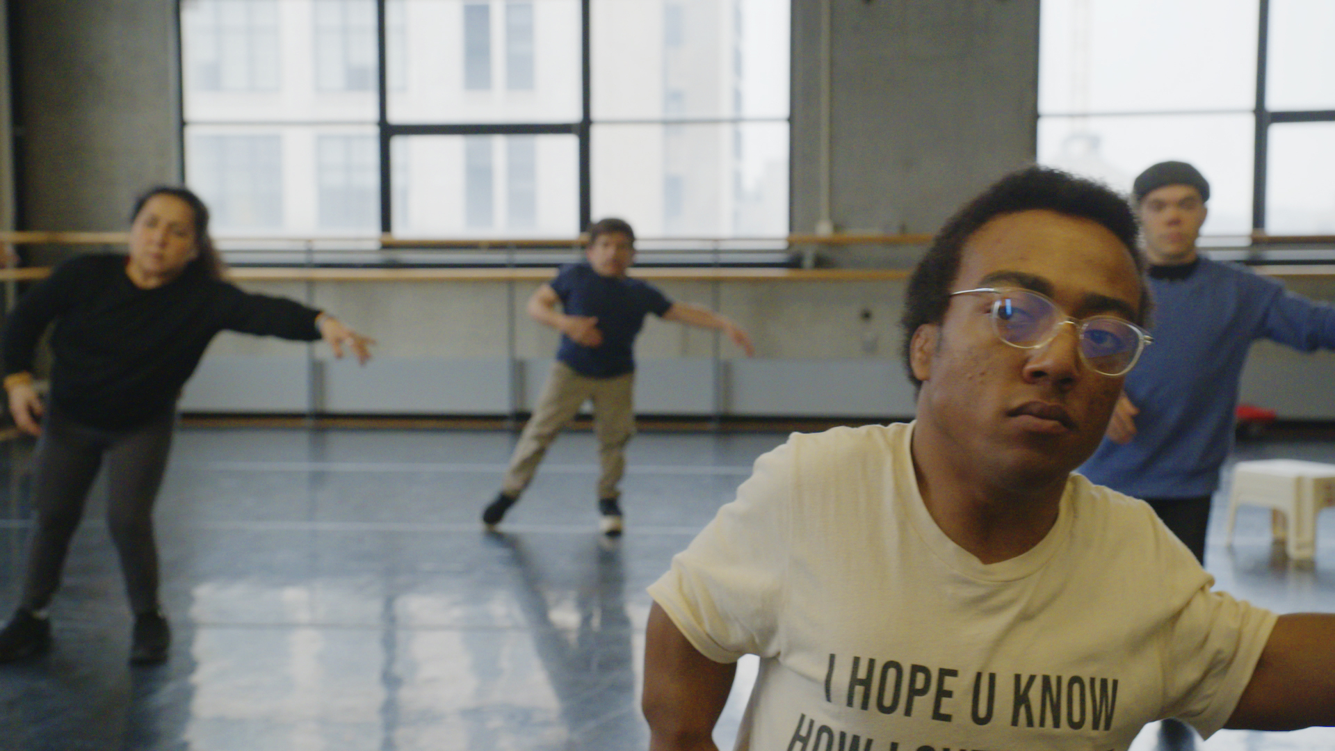 Inside a dance studio, four individuals are learning how to dance with their arm reaching out as they learn the choreography.