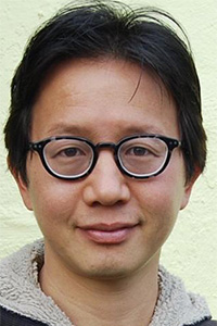 Man with short black hair and glasses smiling against a light-colored wall.
