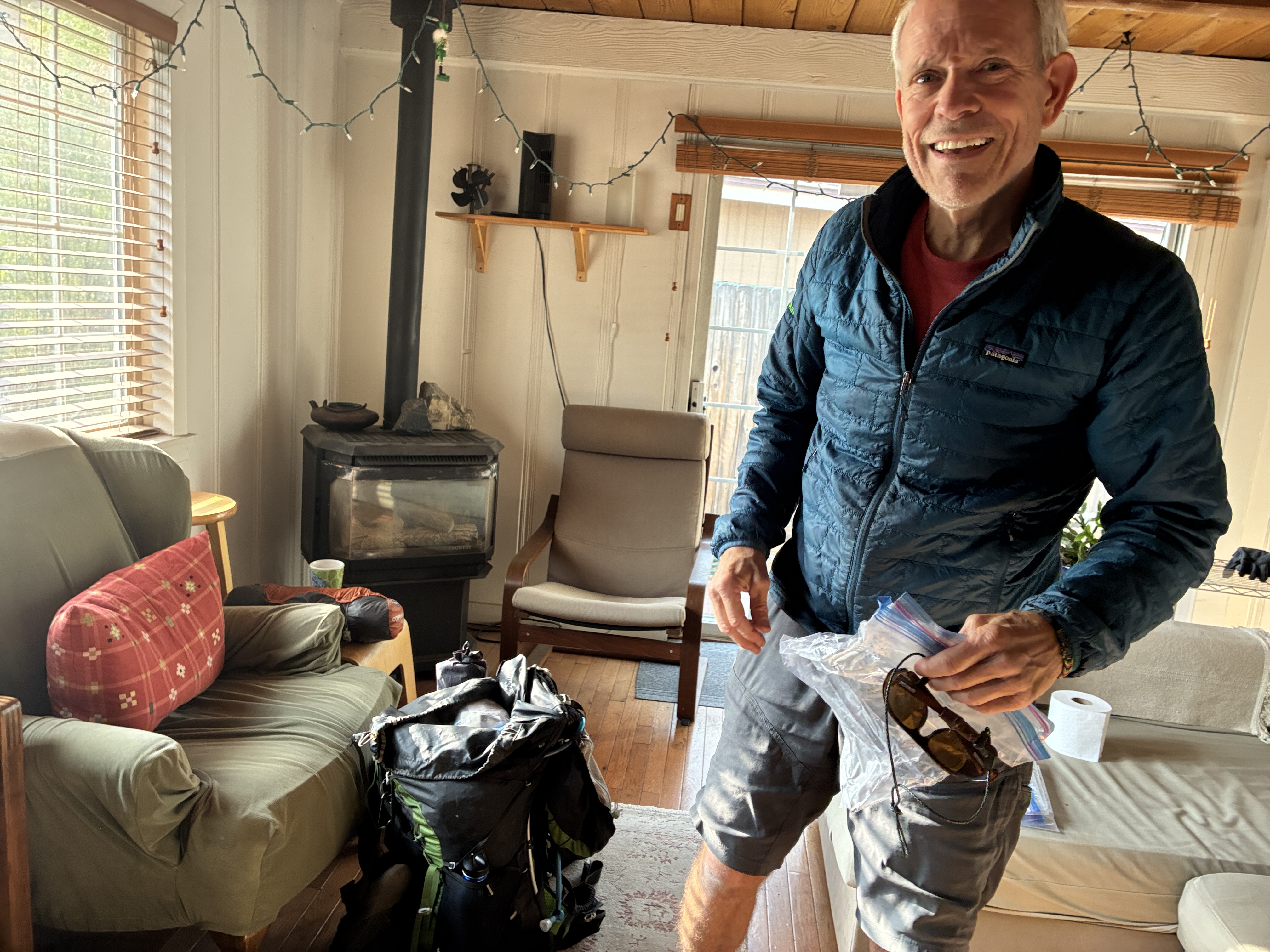 Man in blue puffer jacket stands smiling in a living room while packing for a trip with wood furnace in background