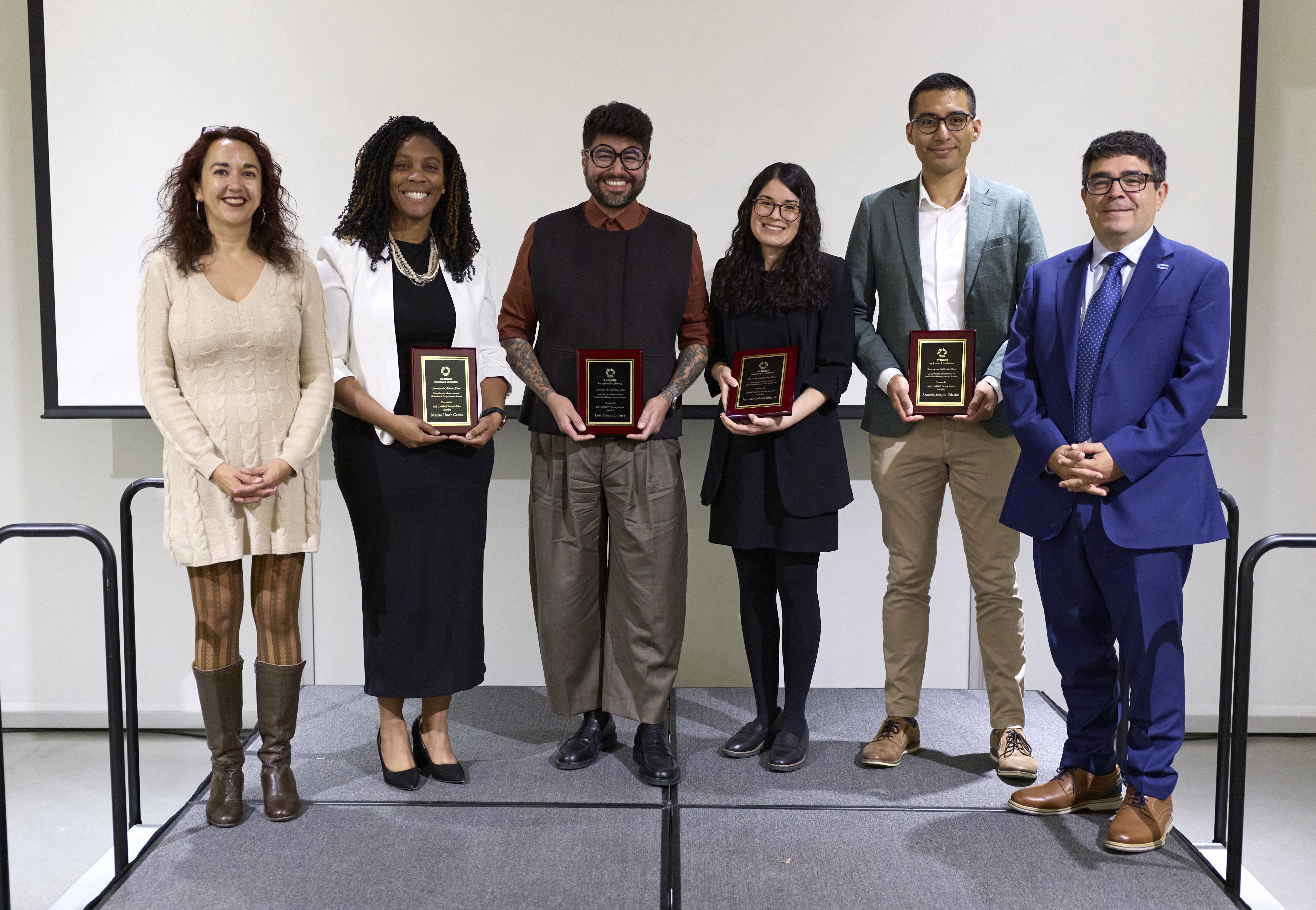 six scholars in professional dress stand atop a stage in front of a blank projection screen. several hold commemorative plaques, with all smiling and facing the camera. 