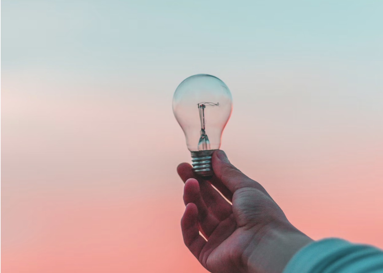 Hand holding clear light bulb against pastel blue-pink sky — photograph