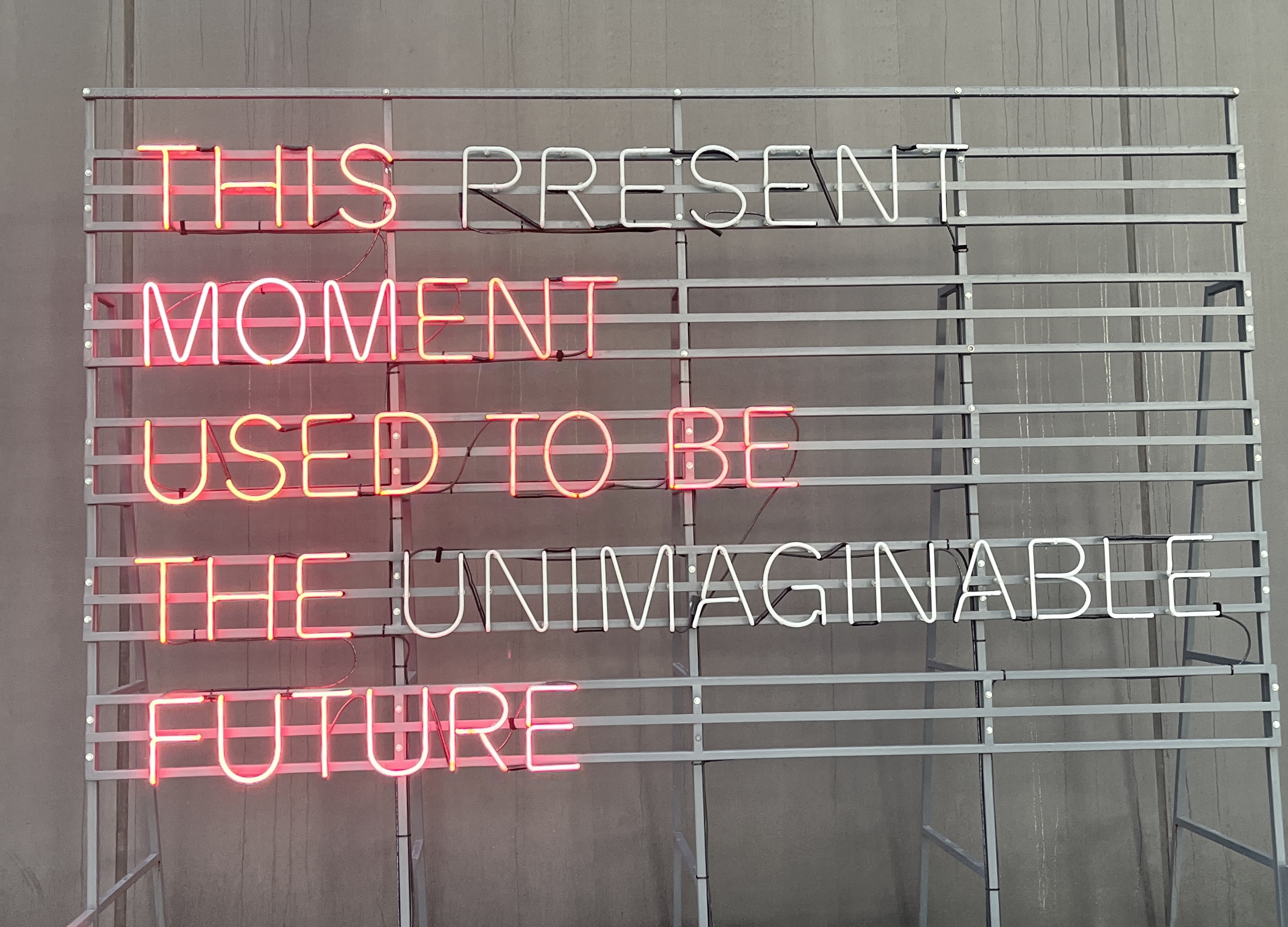 Neon sign with pink letters spelling out “This present moment used to be the unimaginable future.”