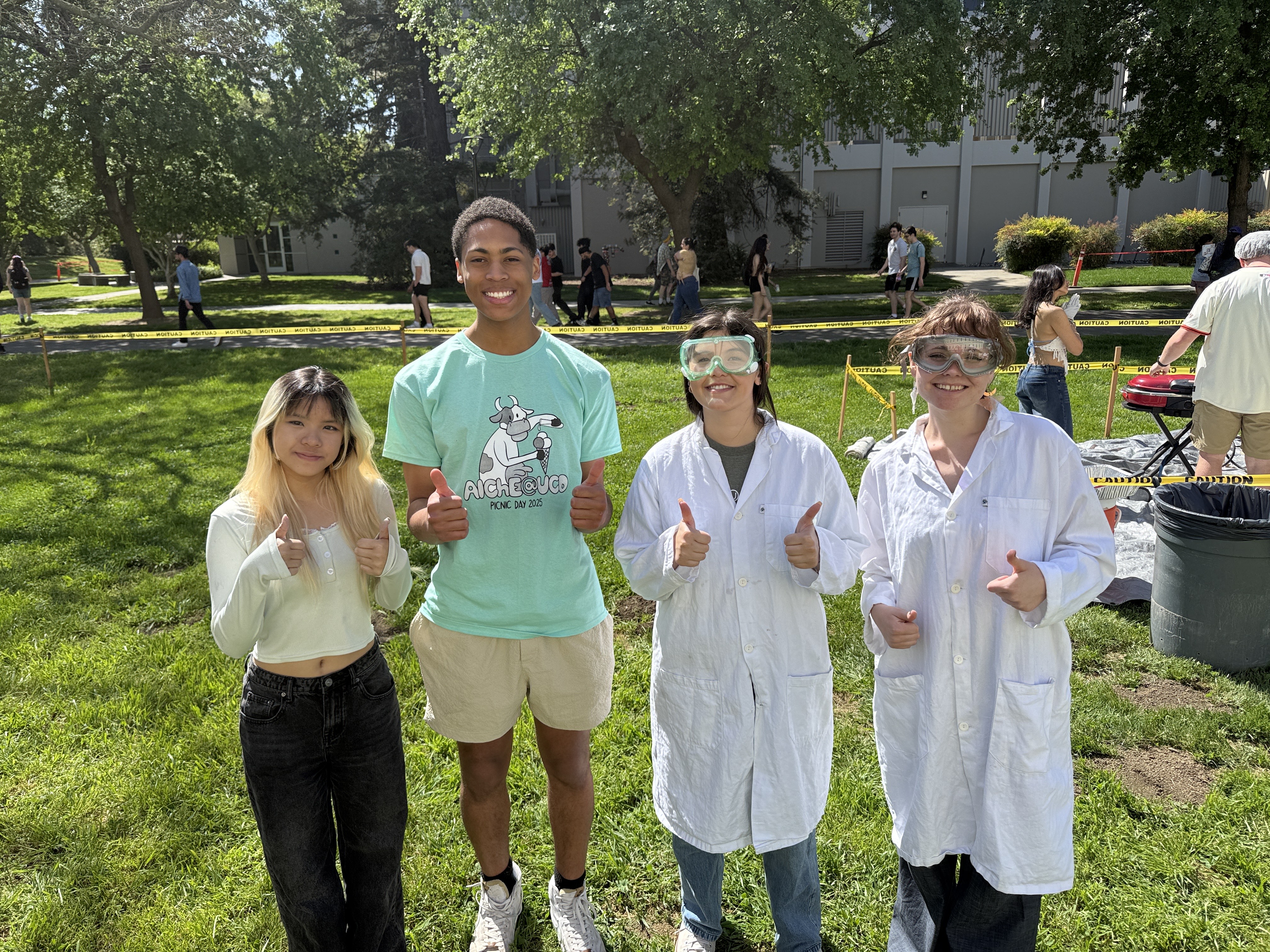 In an outdoor lawn setting, four students stand in a line smiling and giving thumbs up signs to the camera.