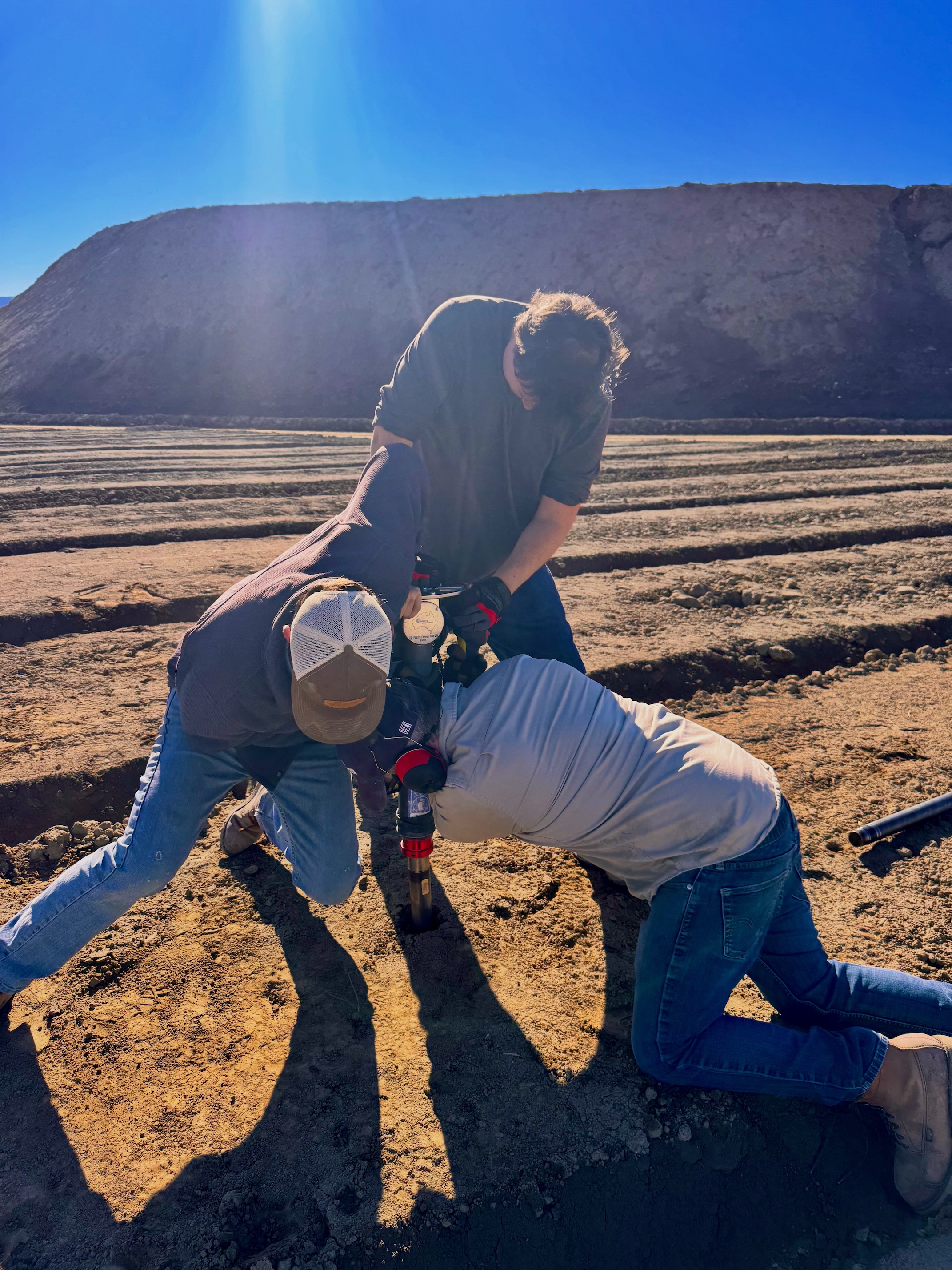 Three researchers bend over farm equipment, one kneeling, while exerting energy, on plowed field with mountain in background under blue sky