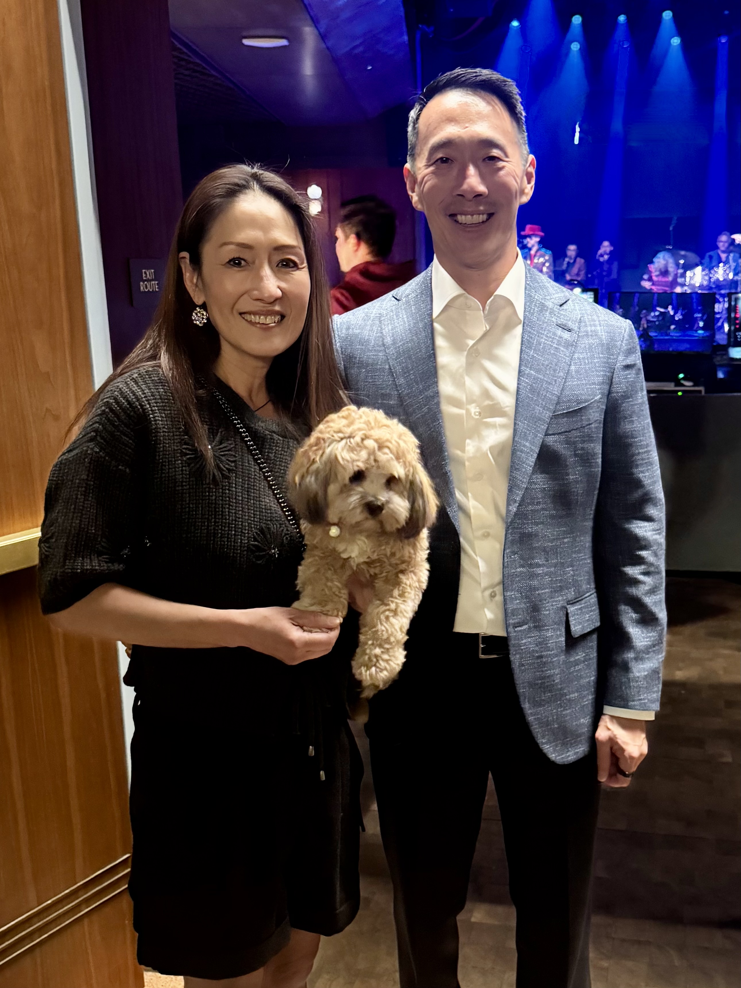Smiling couple at dim venue; woman holds small curly tan dog with blue stage lights behind. Kathy Chiao and Ken Hao and their dog King King. The couple has given the second largest individual gift to the Weill School of Veterinary Medicine. (Ken Hao)