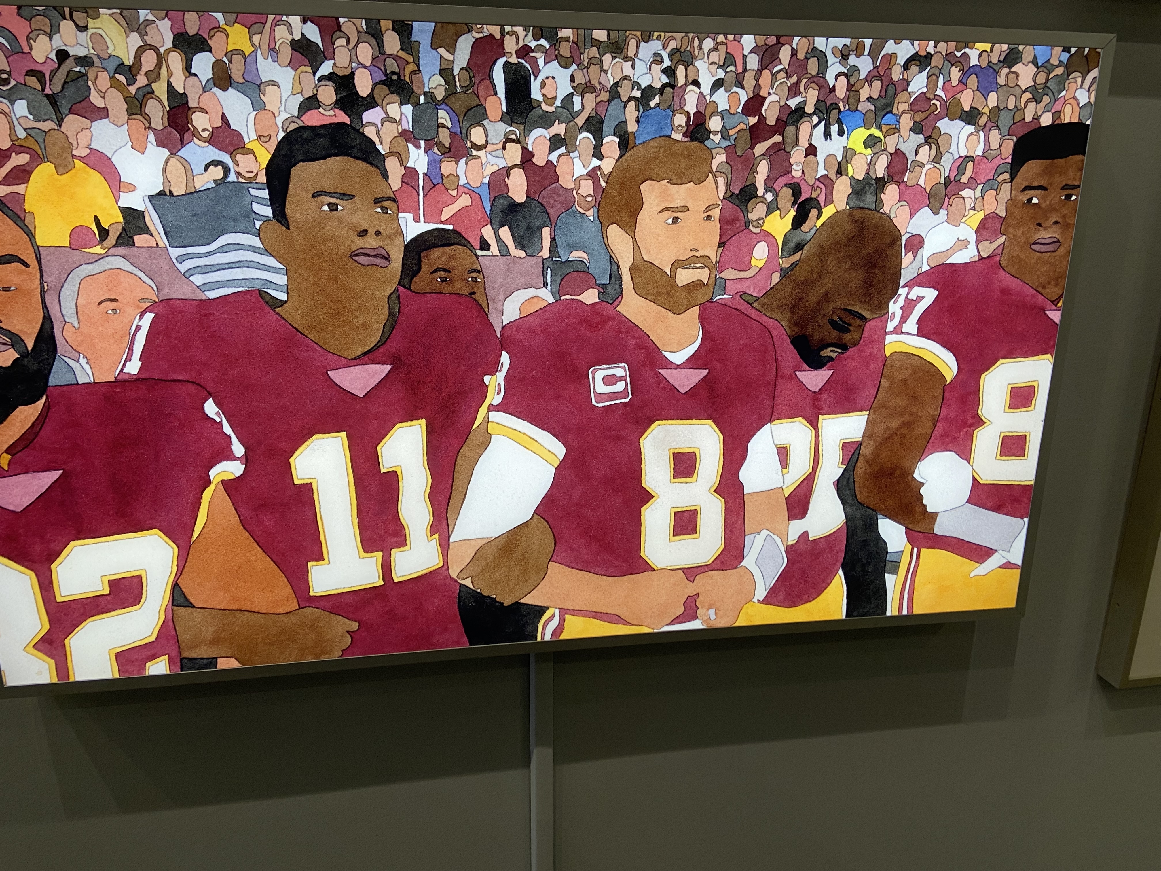 Group of football players in maroon uniforms standing together, with a cheering crowd in the background.