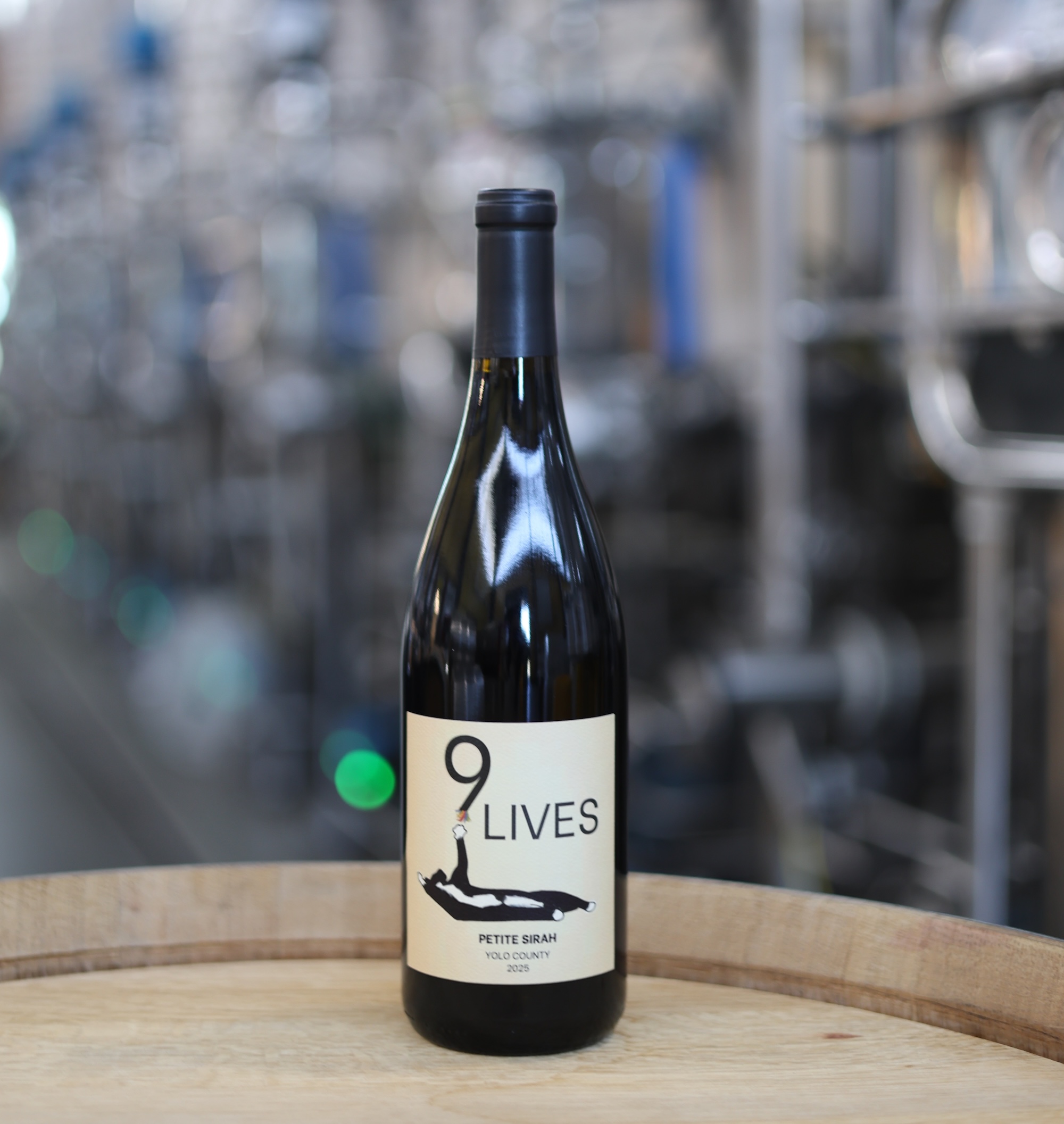 9 Lives wine bottle on wooden barrel with blurred winery tanks in background. The 9 Lives 2025 Yolo County Petite Sirah, one of the student-made wines available for sale. (UC Davis) 