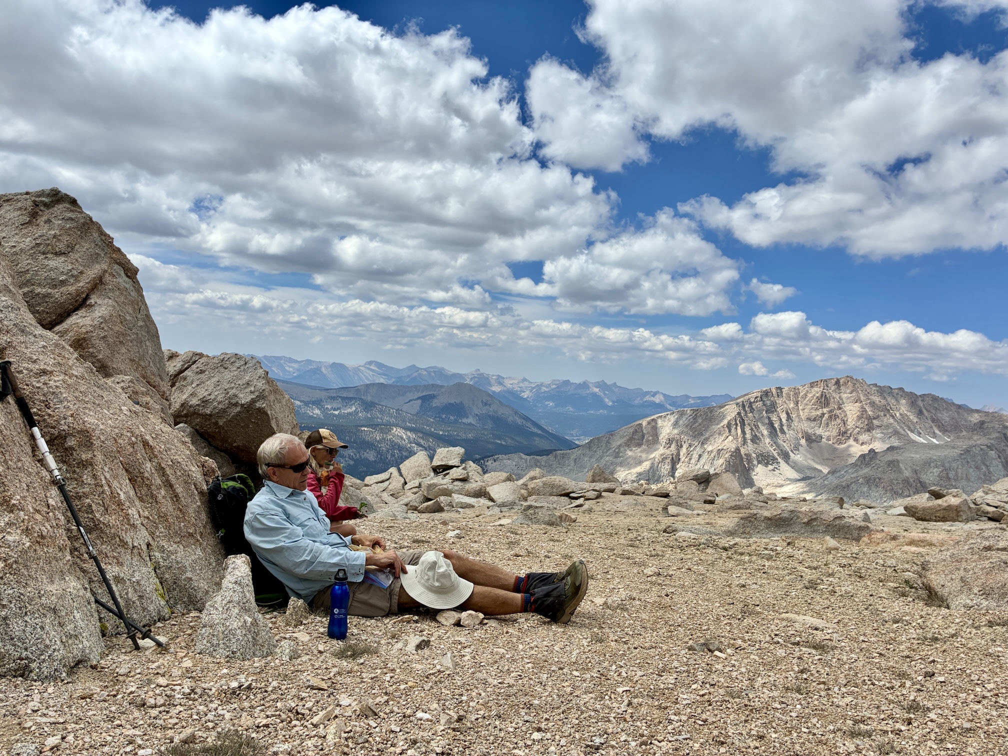 Man in blue shirt and woman in red shirt slump resting against boulder with vista or Sierra mountains in background