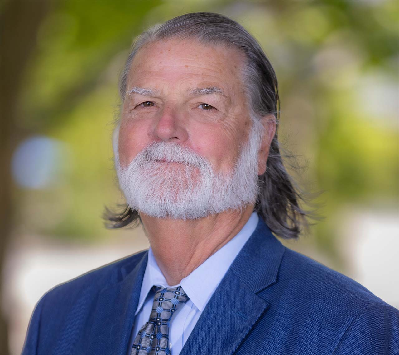 A bearded man in a blue suit smiles, with greenery in the background.